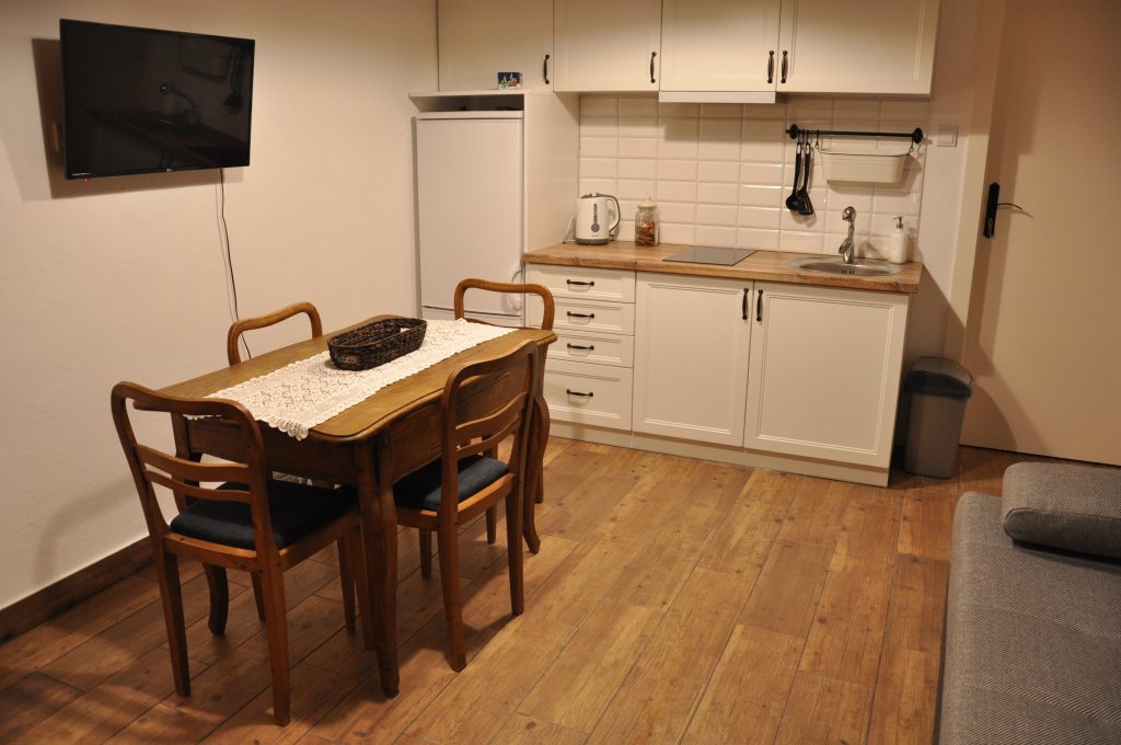 Widok pomieszczenia, na pierwszym planie drewniany stół z czterema krzesłami, za nim białe szafki kuchenne z drewnianym blatem, zlewem i kuchenką, obok lodówka.