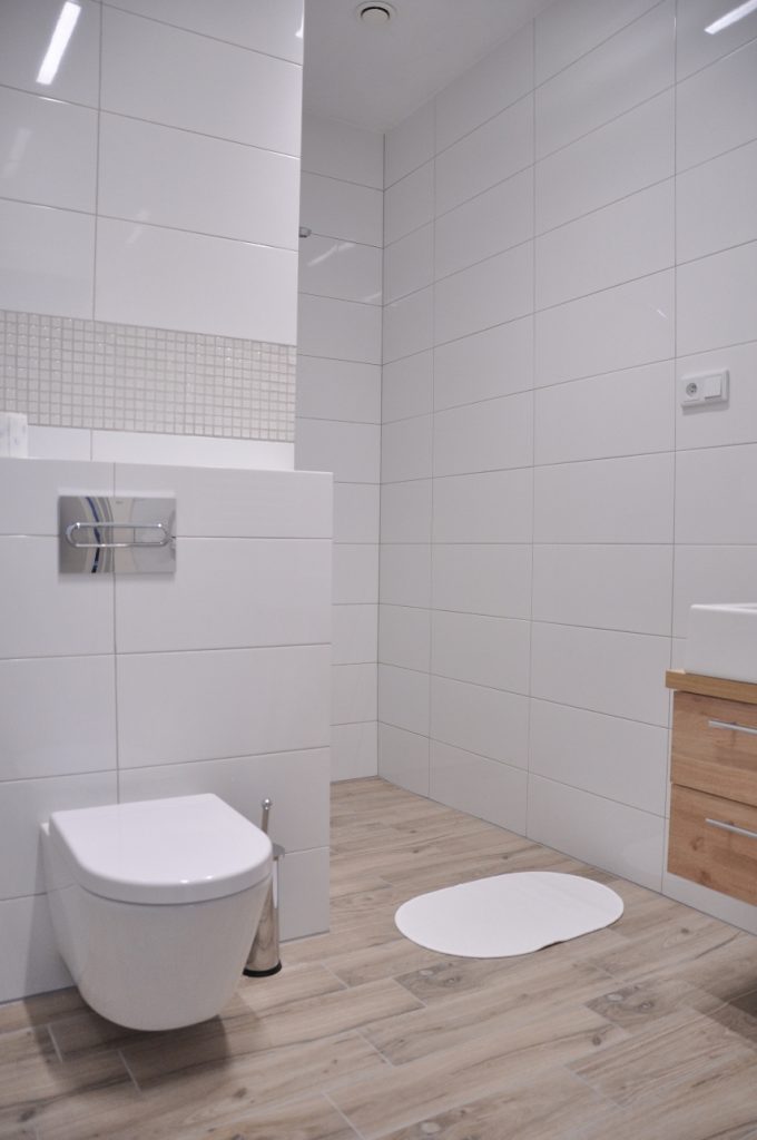 Widok łazienki, z przodu podwieszana toaleta, na ścianach białe płytki, na podłodze szare płytki, na nich biały dywanik. W głębi wnęka prysznicowa